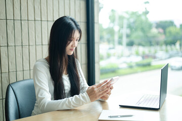 Obraz premium Gen Z Young Woman Using Smartphone at Desk with Laptop in Modern Cafe Office Setting