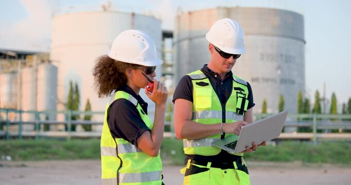 Female refinery technician reporting operational update through walkietalkie while engineer reviews maintenance checklist on laptop near petrochemical storage infrastructure