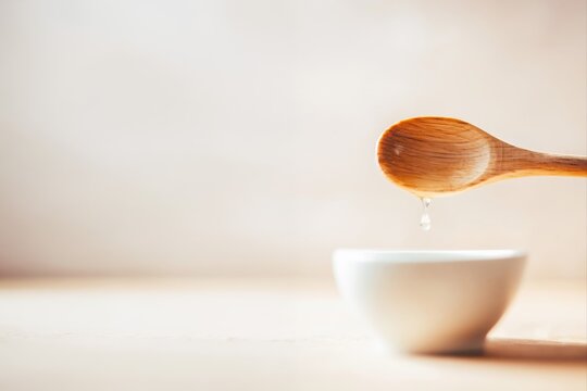 A wooden spoon with a droplet falling into a white bowl on a light background. Minimalist composition highlights natural kitchen utensils and clean food preparation.