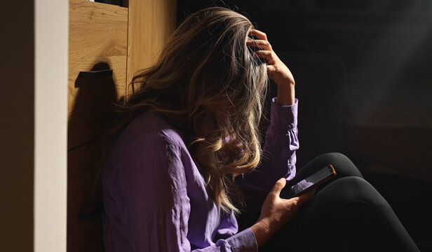 Depressed woman holding smartphone sitting on floor in darkness.