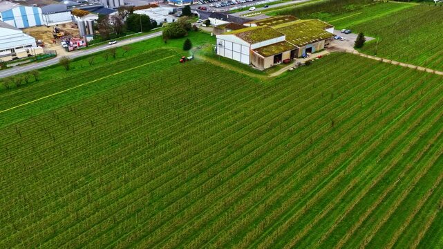 Aerial view of a tractor working in green agricultural fields next to a farm building in Europe.