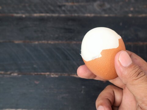 Close up view of hand holding a freshly boiled egg with part of its shell peeled off on dark table background. Ideal for concepts related to healthy breakfast, protein snack, cooking, and nutrition