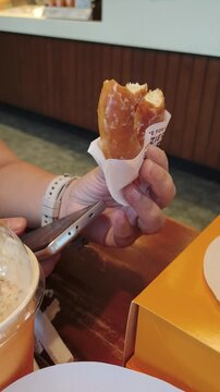 Hand holding half-eaten glazed doughnut with drink beside on wooden table