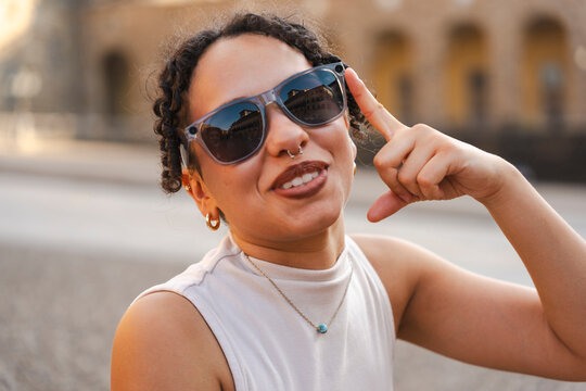 A happy young woman with curly hair smiles outdoors, wearing sunglasses while making a phone call hand gesture during sunset in a historic European city.