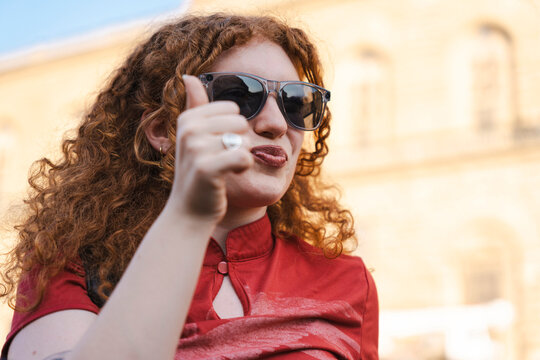 A young woman with curly red hair and sunglasses gives a thumbs up gesture while making a playful face. She wears a red shirt with a blurred city background.