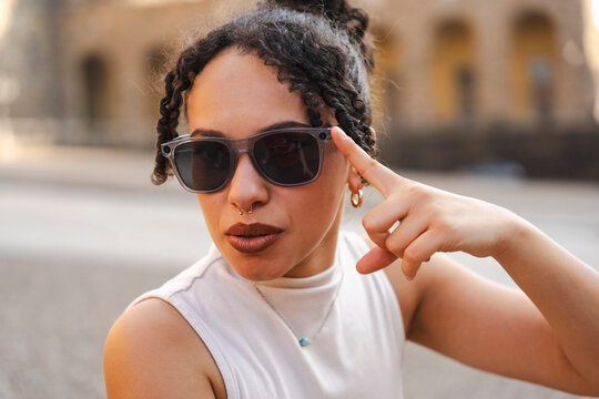A stylish young woman with curly hair and a septum piercing points to her sunglasses. She looks directly at the camera while posing in a sunny outdoor setting.