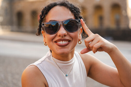 A cheerful young woman with curly braided hair and stylish sunglasses smiles warmly at the camera while making a playful hand gesture on a sunny city street.