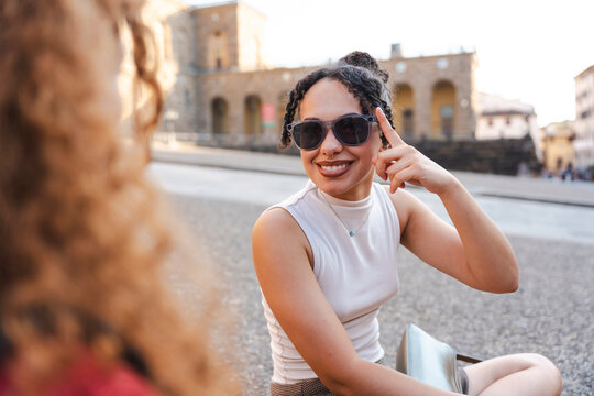 A smiling young woman wearing sunglasses sits outdoors on a stone plaza, chatting and gesturing happily while spending some quality time with her female friend.