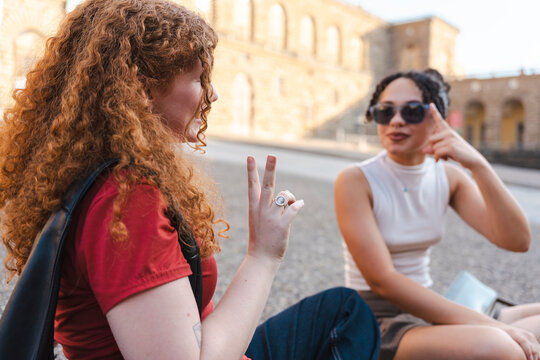 Two young diverse female friends, one with curly red hair, communicate using sign language while sitting together outdoors in a sunny historic European plaza.
