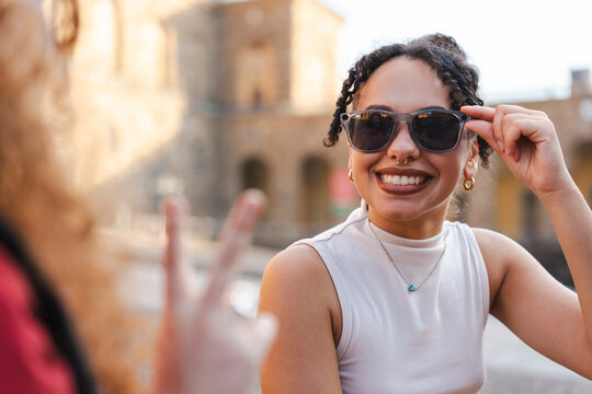 A smiling young woman wearing sunglasses and a septum ring enjoys a sunny day outdoors with a friend who shows a peace sign, set against a historic European city background.