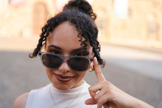 A confident young woman with curly black hair smiles at the camera, lowering her stylish sunglasses and gesturing with her index finger on a sunny city street.