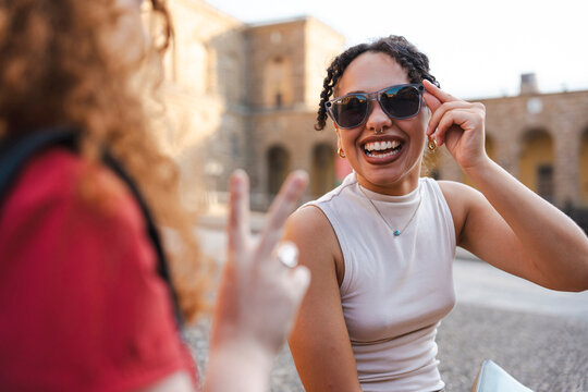 Two young diverse female friends laugh together in a historic city plaza during summer. A woman wearing sunglasses smiles as her friend makes a peace sign.