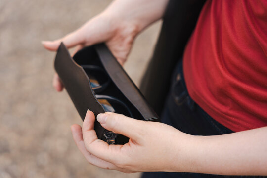 Close-up of a woman's hands opening a dark brown leather glasses case to reveal eyeglasses inside. She is wearing a vibrant red shirt and a dark jacket outdoors.