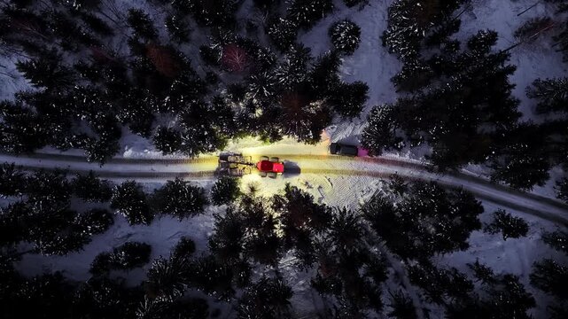 Aerial view of a snow groomer with bright headlights clearing a snow-covered forest road at night, surrounded by dark pine trees.