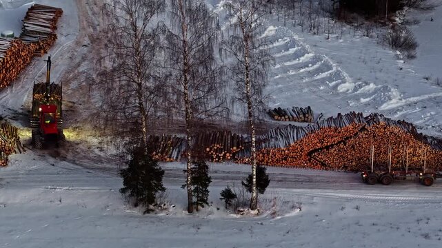 Aerial view of a red forestry machine working among large stacks of timber in a snow-covered forest landscape in Europe.