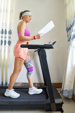 Athletic woman with knee brace exercising on treadmill home while holding paper documents for multitasking indoor workout training.