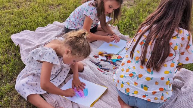 A group of children enjoy drawing on paper with crayons while sitting on a blanket in a grassy area. It is a sunny afternoon and they are focused on their art