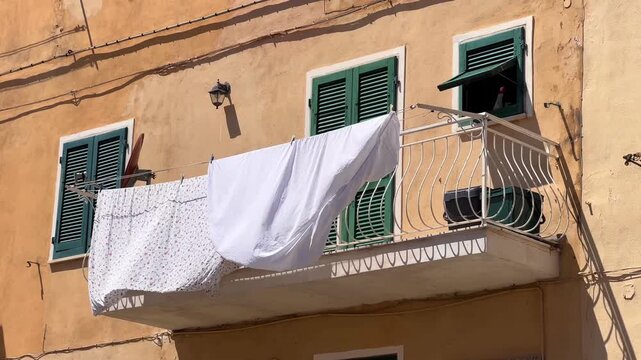 White bedsheets drying on a balcony of an old building with faded yellow colored walls and green shutters in a traditional Italian town authentic lifestyle.