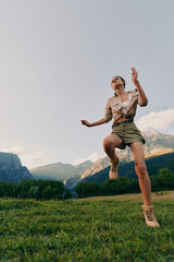 Obraz premium Woman jumping in a meadow with mountains in the background, casual outdoor attire, bright sky, and grass underfoot, capturing a vibrant lifestyle and energetic outdoor moment.