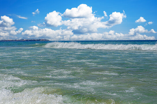 Foamy ocean waves roll towards the shore under a bright blue sky filled with puffy white clouds.