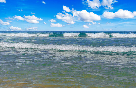 Gentle sea waves roll onto a sunny beach under a bright blue sky filled with white clouds.
