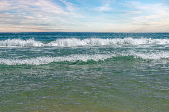Ocean waves crash along the shore under a partly cloudy sky during a bright day.