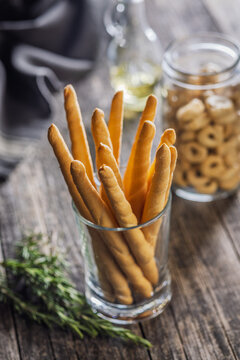 Crunchy Italian Breadsticks (Grissini) In Glass With Herbs, Olive Oil And Jarred Snacks On Wooden Table