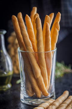 Crunchy Italian Breadsticks (Grissini) In Glass With Herbs, Olive Oil And Jarred Snacks On Wooden Table
