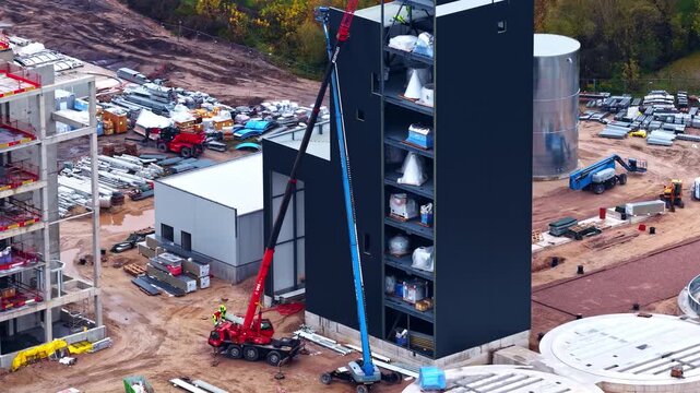 Aerial view of a construction site featuring a tall black industrial tower, a large silver storage tank, and circular concrete foundations in Europe.