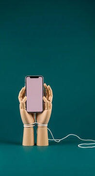 Wooden mannequin hands hold a phone, bound by a cord, symbolizing addiction or entrapment with technology, perfect for digital detox concepts.