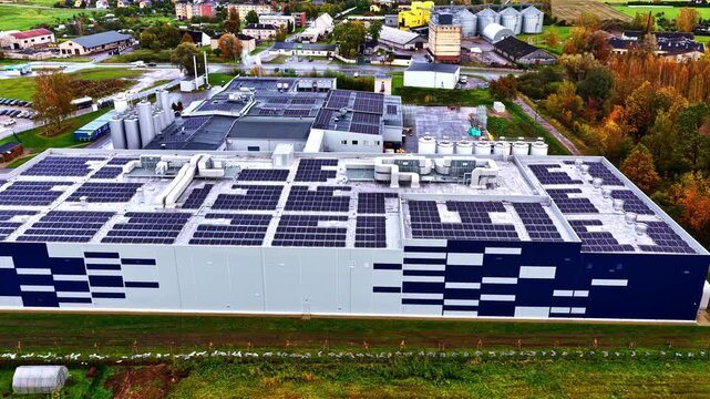 Aerial view of a large industrial facility with extensive solar panel arrays on its flat roof, surrounded by green fields and rural landscape.