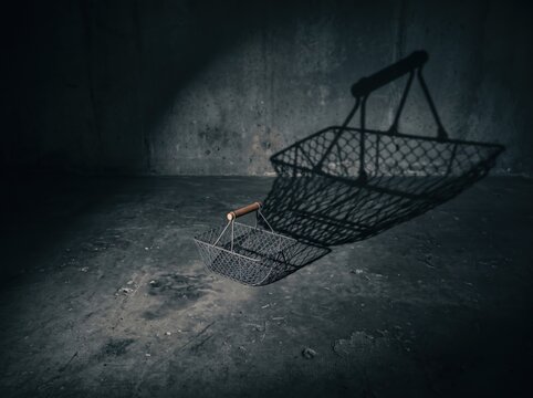 Empty Shopping Cart in Darkness: An abandoned, rusty shopping cart sits isolated on a gritty concrete floor, casting a long shadow in a dimly lit, industrial space.