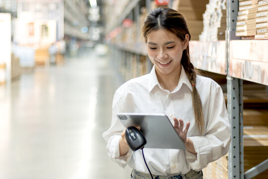 Asian woman worker holds tablet and laser barcode reader counting and checking inventory on shelves in a wholesale warehouse. Young beautiful female logistic staff checking stock in warehouse store.