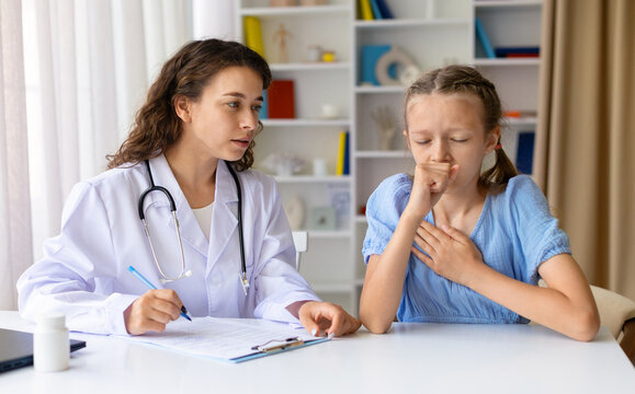 Young girl coughing into her hand during pediatric consultation while doctor taking notes at a desk in clinic setting, illustrating respiratory symptoms and professional medical care