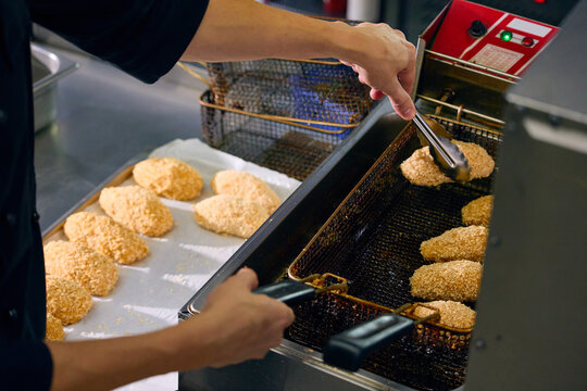Professional cook transferring breaded food portions beside deep fryer in commercial kitchen. Concept of restaurant food preparation, commercial kitchen workflow, catering production.