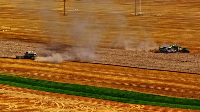 Aerial view of combine harvesters working in a golden grain field, creating dust clouds while harvesting crops in Europe.