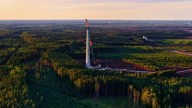 Aerial view of a wind turbine tower under construction surrounded by dense pine forest during golden hour in Europe.
