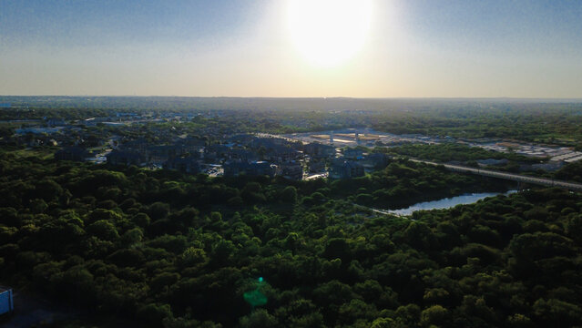 Bright sun over woodland, intense solar glare illuminates dense forest canopy along Leon Creek in Northwest San Antonio, Texas. Distant multifamily housing complexes border horizon line
