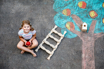 Naklejka premium Cute little girl painting with colorful chalks apples harvest from apple tree on asphalt. Cute preschool child with having fun with chalk picture. Creative leisure for children, drawing and painting.