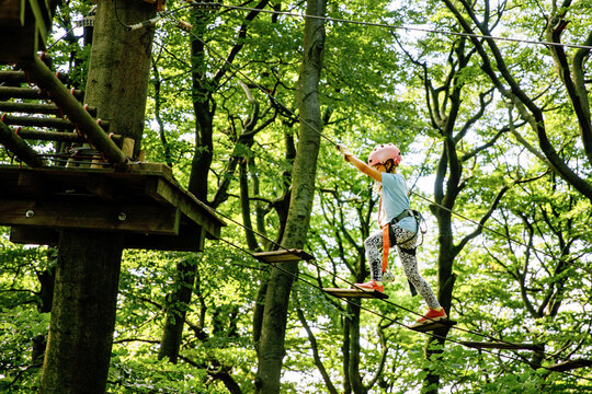 Happy little girl climbing rope course in forest adventure park, child wearing helmet and harness enjoying outdoor activity, real life moment. School child having fun.
