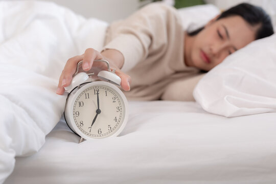 Tired Asian woman in comfortable bed reaching hand to snooze white alarm clock during early morning showing sleepy reluctant expression about waking up inside cozy bedroom.