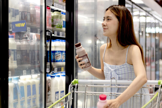 Asian woman picking milk bottle from refrigerator during pushes a shopping cart down an aisle to buy food and beverage in supermarket. Beautiful customer select milk and dairy products in store fridge