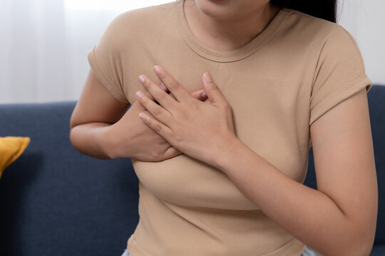 Uncomfortable distressed woman holds hand on chest feeling sudden pain and health problem sitting on sofa experiencing severe breast discomfort and acute medical symptom needing urgent attention