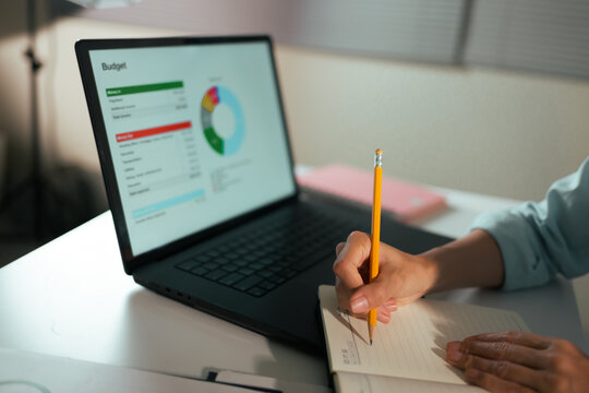 Person's hands writing in a notebook while analyzing financial data on a laptop screen, managing personal or business budget