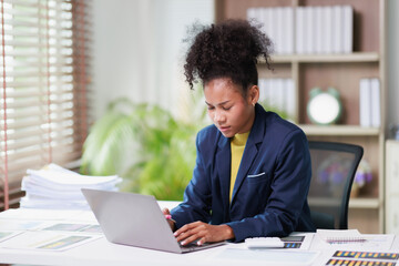 Fototapeta premium Young black businesswoman working intensely on her laptop at an office desk, focused on tasks, possibly feeling the pressure of a heavy workload and deadlines