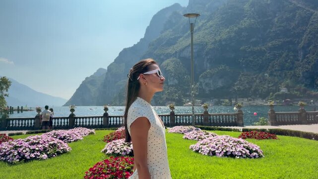 Elegant Woman Walking in Flower Garden by Lake Garda, Scenic Riva del Garda Waterfront with Colorful Flowers and Alpine Mountains, Summer Travel Lifestyle in Trentino Alto Adige, Italy