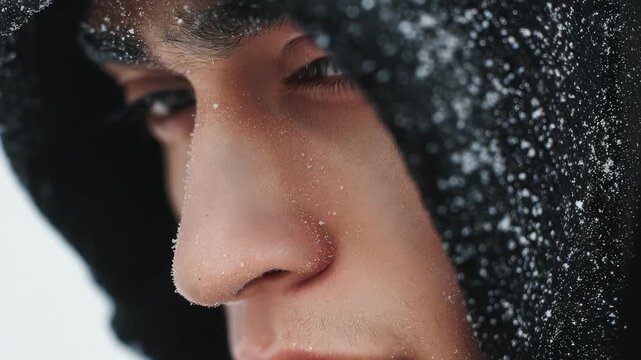 A detailed close up of a male face partially hidden by a hood, with snowflakes resting on the fabric and skin. The image captures a sense of winter chill and enigmatic emotion