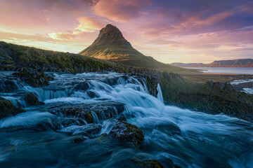Silky waterfall cascading over mossy rocks below an iconic cone shaped mountain under a pink sunset sky © Kafi