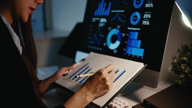 Young professional woman working late, analyzing graphs and charts on a computer screen and paper documents in a dimly lit office