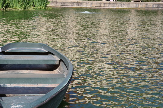 Empty green rowboat floating on a calm garden pond with rippling reflections, surrounded by lush aquatic plants and a stone embankment in daylight.